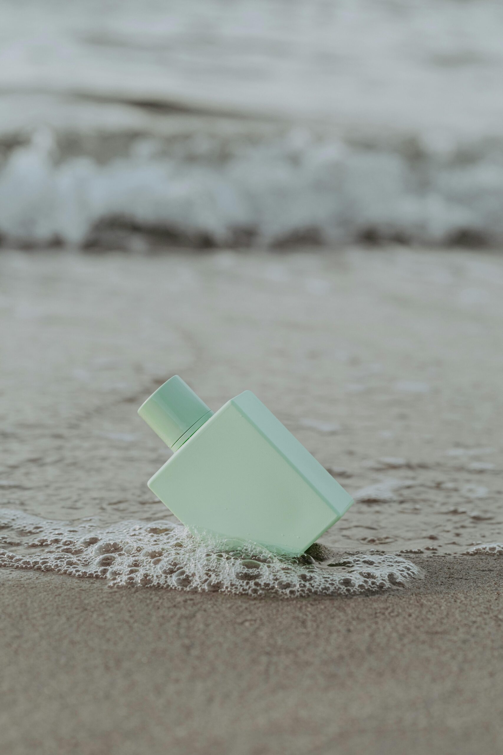Minimalist perfume bottle resting on sandy beach with gentle sea foam.