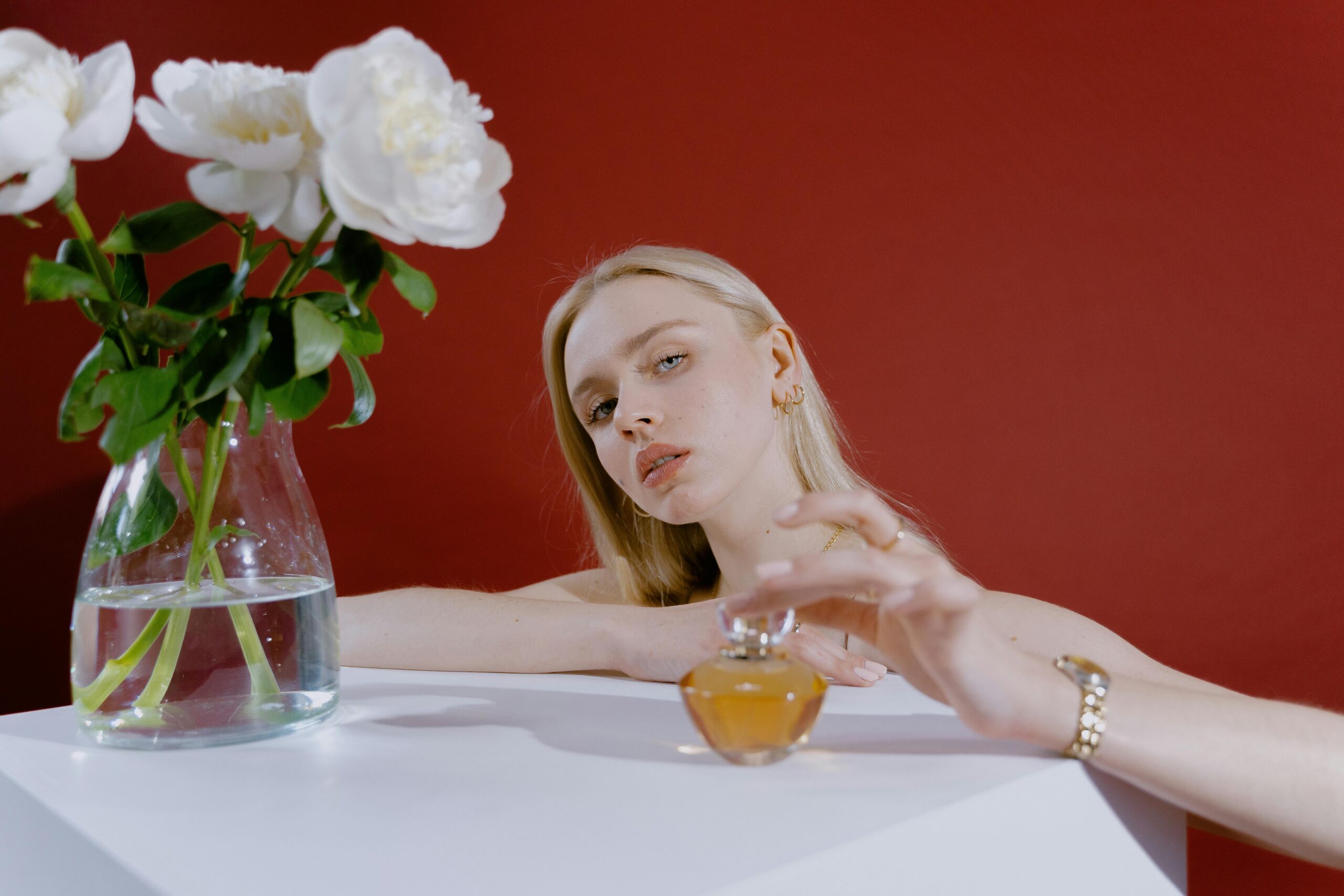 A woman with blond hair and a perfume bottle next to a vase of white roses on a white table.